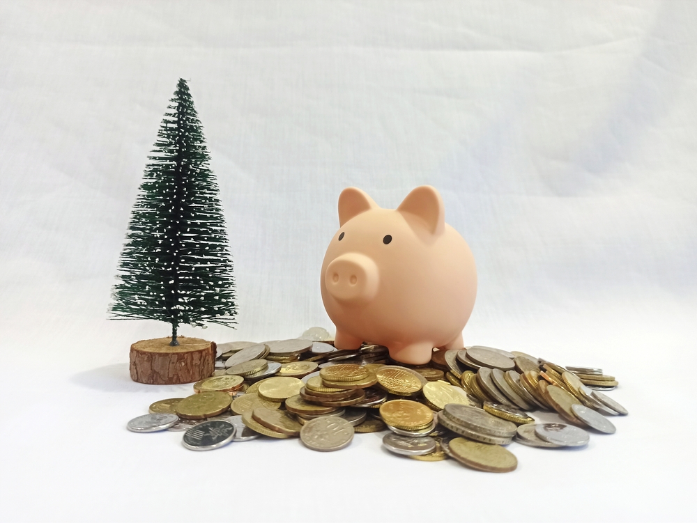 Pink piggy bank surrounded by coins next to a small artificial Christmas tree, symbolizing financial savings, holiday budgeting expenses management